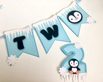 Pode incluir: Uma faixa azul e branca com tema de pinguim com a palavra "DOIS" e um grande número "2" com um pinguim nela. A faixa tem flocos de neve brancos e pingentes de gelo.