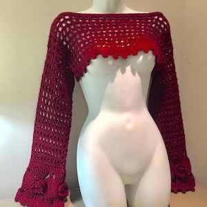 May include: A burgundy crochet shrug with long sleeves and a scalloped neckline. The sleeves feature floral accents near the cuffs. The garment is displayed on a white mannequin.