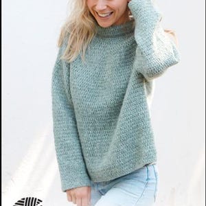 May include: A light blue, textured knit sweater with long sleeves. The sweater is worn by a person with blonde hair, smiling. The image includes the text "DROPS DESIGN" and the website address www.garnstudio.com.