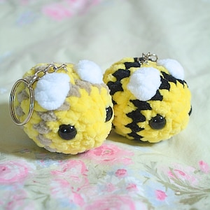 May include: Two handmade, plush bee keychains. The bees are yellow with black stripes and white accents. Each keychain has a silver keyring and black plastic eyes. The keychains are on a floral background.