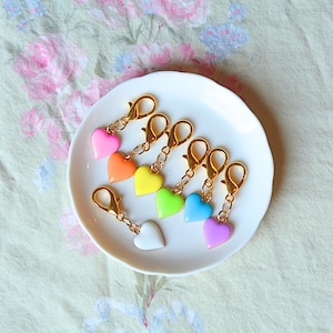 May include: A collection of colorful heart-shaped charms with gold-colored clasps, arranged on a white plate. The charms are in various colors, including pink, orange, yellow, green, blue, white, and purple. The background is a floral patterned fabric.