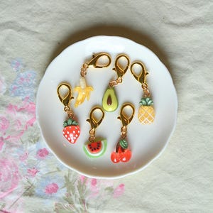 May include: A collection of fruit-themed charms with gold-colored clasps. The charms include a strawberry, banana, avocado, watermelon, cherry, and pineapple. The charms are arranged on a white plate, set against a floral patterned background.