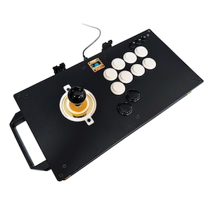 May include: A black arcade controller featuring a central joystick with a black ball top, eight white action buttons, two black buttons, and a small digital display screen. The rectangular device includes a side handle and a wired connection.
