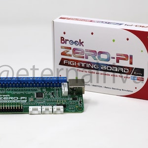 Puede incluir: Una placa de circuito verde Brook Zero-Pi Fighting Board con bloques de terminales azules y conectores blancos, colocada junto a su caja de venta blanca y roja etiquetada Brook Zero-Pi Fighting Board con texto sobre mandos arcade.