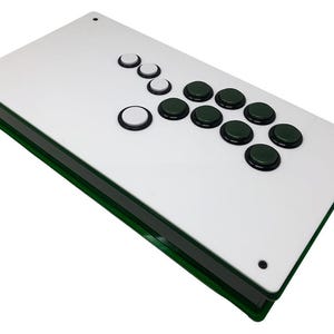 May include: A rectangular arcade controller featuring a white top panel with a green side frame. The layout includes three white circular buttons and eight dark green circular buttons arranged in a standard gaming configuration on the surface.