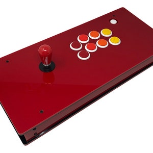 Könnte beinhalten: Ein roter Arcade-Gamecontroller mit einem Joystick und acht farbigen Tasten. Der Controller hat eine rechteckige Form und eine glänzende Oberfläche. Der Joystick hat einen roten Knauf.
