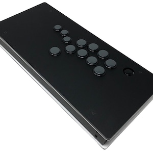 Op de afbeelding: Een zwarte arcade game controller met een rechthoekig ontwerp. Het heeft een reeks grijze knoppen gerangschikt in een directioneel pad en actieknop layout. De controller staat tegen een witte achtergrond.