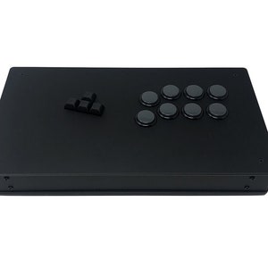 Könnte beinhalten: Ein schwarzer Arcade-Stick-Controller mit acht runden Knöpfen und einem Richtungspad. Der rechteckige Controller hat eine matte Oberfläche. Abmessungen: ca. 30 cm x 20 cm.
