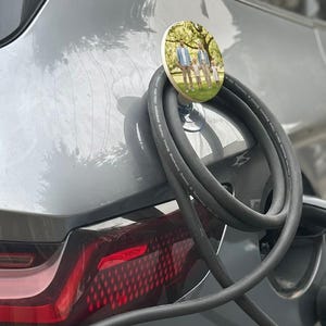 Könnte beinhalten: Ein runder Kabelhalter für ein Autoladegerät aus Holz mit einem Familienfoto, das an einem grauen Auto befestigt ist. Der Halter wird mit einem Saugnapf am Auto befestigt. Ein schwarzes Ladekabel ist mit dem Auto verbunden.