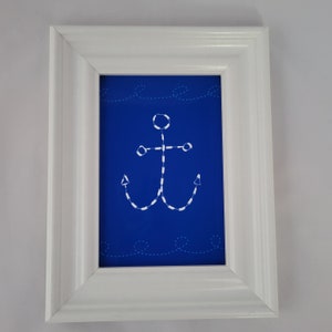 May include: A white framed picture with a blue background and a white anchor with a dotted line design. The anchor is surrounded by white dotted lines that resemble waves.