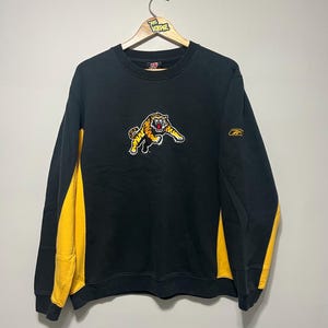May include: Black sweatshirt with yellow side panels and a tiger graphic. The tiger is yellow, orange, and white. The sweatshirt has a crew neck and long sleeves. A small Reebok logo is on the right sleeve.