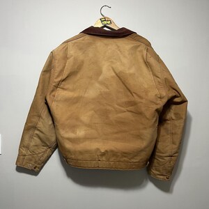 Puede incluir: Una chaqueta de trabajo marr&oacute;n con un cuello de pana marr&oacute;n cuelga de una percha de madera. La chaqueta tiene un forro acolchado y pu&ntilde;os abotonados. La parte trasera de la chaqueta es visible, mostrando las costuras y la forma general.