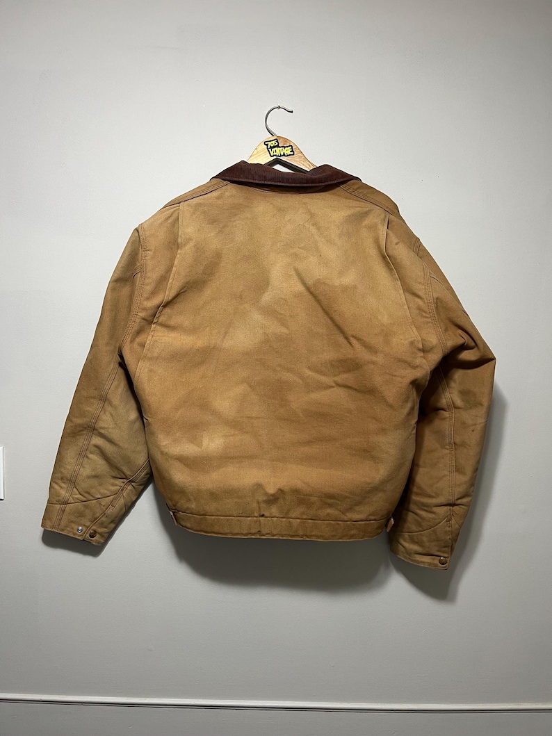 Puede incluir: Una chaqueta de trabajo marr&oacute;n con un cuello de pana marr&oacute;n cuelga de una percha de madera. La chaqueta tiene un forro acolchado y pu&ntilde;os abotonados. La parte trasera de la chaqueta es visible, mostrando las costuras y la forma general.