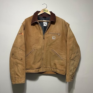 Puede incluir: Una chaqueta de trabajo Carhartt marr&oacute;n con cuello de pana marr&oacute;n, cremallera y m&uacute;ltiples bolsillos. La chaqueta est&aacute; colgada de una percha de madera. El logotipo de Carhartt es visible en el bolsillo del pecho.