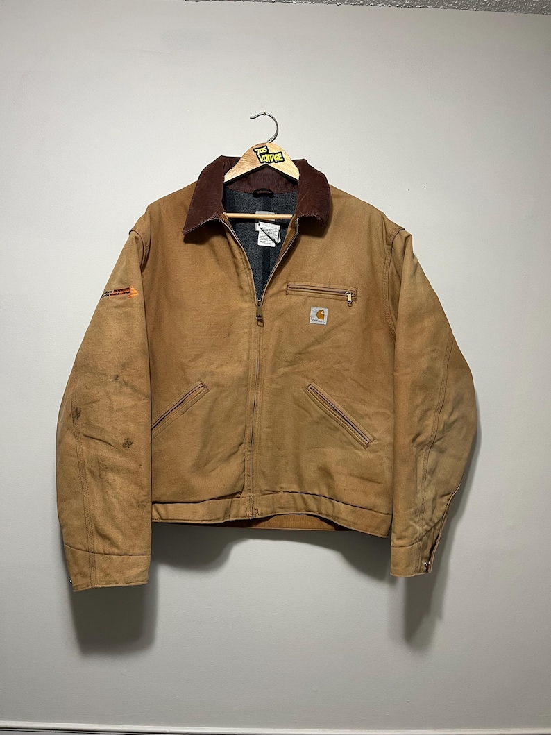 Puede incluir: Una chaqueta de trabajo Carhartt marr&oacute;n con cuello de pana marr&oacute;n, cremallera y m&uacute;ltiples bolsillos. La chaqueta est&aacute; colgada de una percha de madera. El logotipo de Carhartt es visible en el bolsillo del pecho.