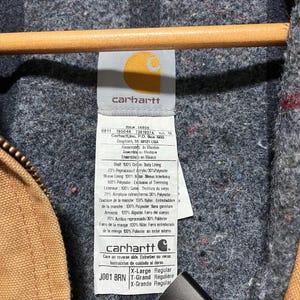 Puede incluir: Primer plano de la etiqueta de una chaqueta Carhartt. La etiqueta muestra el logotipo de Carhartt, la talla y la composici&oacute;n del material. La chaqueta es marr&oacute;n con un forro gris. La etiqueta dice "X-Large Regular".