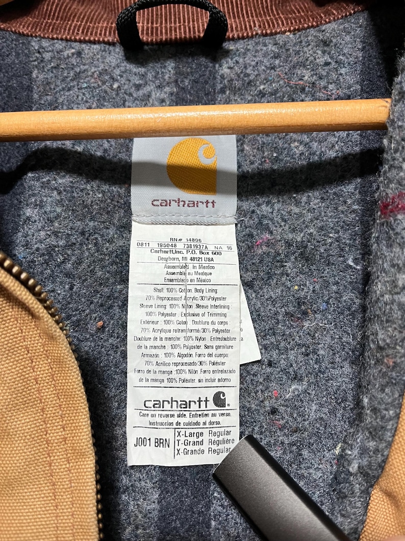 Puede incluir: Primer plano de la etiqueta de una chaqueta Carhartt. La etiqueta muestra el logotipo de Carhartt, la talla y la composici&oacute;n del material. La chaqueta es marr&oacute;n con un forro gris. La etiqueta dice "X-Large Regular".