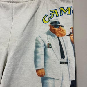 Pu&ograve; includere: Pantaloncini bianchi con una grafica della mascotte delle sigarette Camel in abito bianco, cravatta nera e occhiali da sole. La parola "CAMEL" &egrave; stampata in giallo e blu sopra l'immagine. I pantaloncini hanno un elastico in vita.