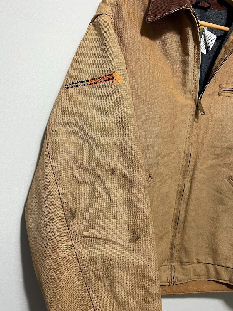 Puede incluir: Chaqueta de trabajo color canela con cuello de pana marr&oacute;n y cremallera completa. La chaqueta tiene un peque&ntilde;o logotipo bordado en la manga izquierda y algunas manchas. La chaqueta est&aacute; hecha de un material duradero y resistente.