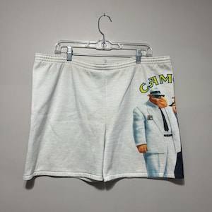 Pu&ograve; includere: Pantaloncini bianchi con elastico in vita con una grafica di un personaggio dei cartoni animati in abito e cappello bianchi. La parola "CAM" &egrave; stampata in giallo e verde sui pantaloncini. I pantaloncini sono appesi a una gruccia di plastica trasparente.