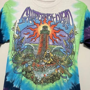 May include: Tie-dye t-shirt featuring a colorful graphic of a lighthouse, skeleton in a boat, and the words "Grateful Dead" in a stylized font. The shirt has a white base with blue, green, and purple tie-dye patterns on the sleeves and sides.
