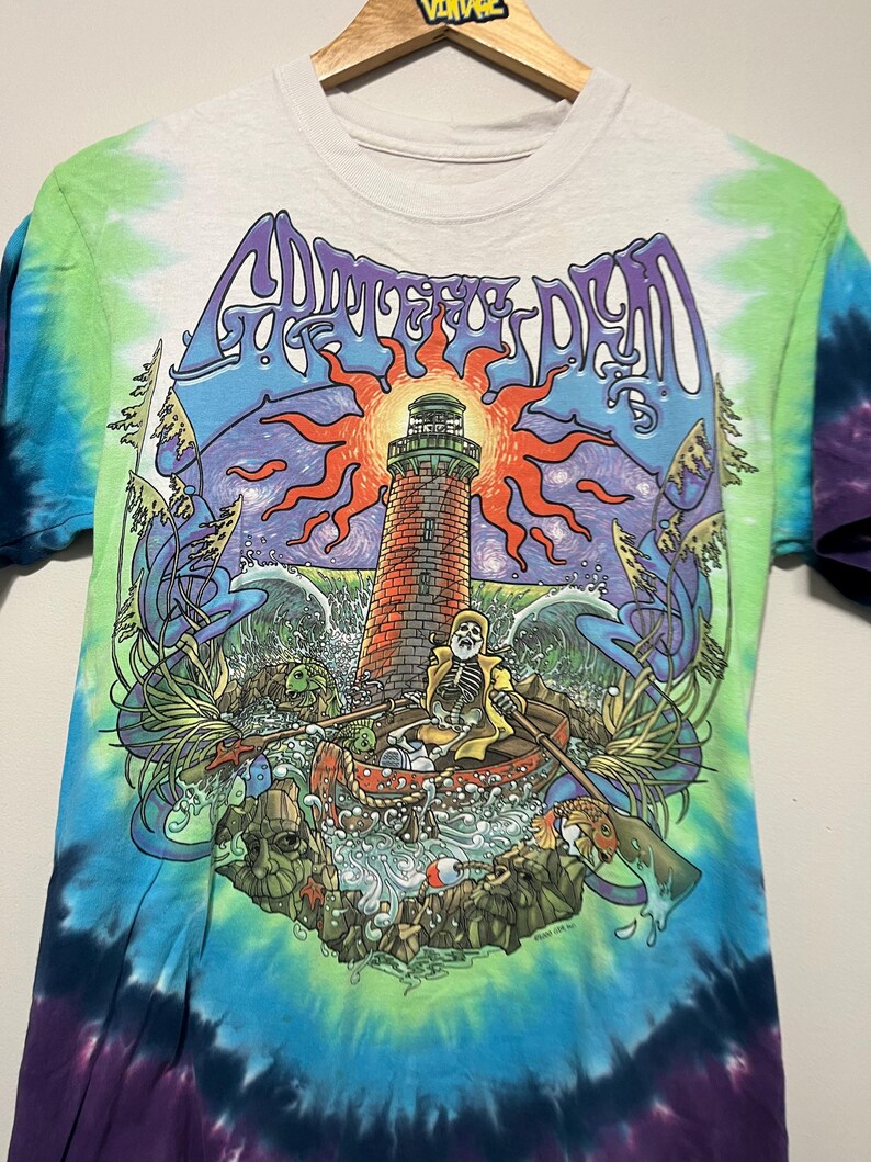 May include: Tie-dye t-shirt featuring a colorful graphic of a lighthouse, skeleton in a boat, and the words "Grateful Dead" in a stylized font. The shirt has a white base with blue, green, and purple tie-dye patterns on the sleeves and sides.