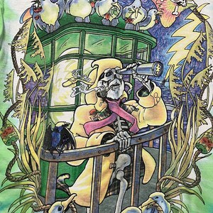 May include: Tie-dye t-shirt featuring a colorful illustration of a skeleton in a yellow coat looking through a telescope. The design includes cartoon birds, a crow, and a skull with a tie-dye brain.