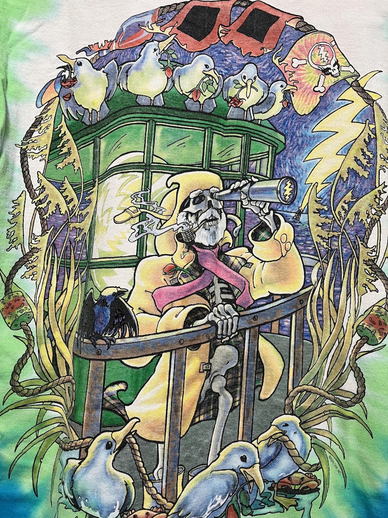 May include: Tie-dye t-shirt featuring a colorful illustration of a skeleton in a yellow coat looking through a telescope. The design includes cartoon birds, a crow, and a skull with a tie-dye brain.