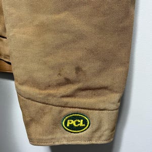 Puede incluir: Primer plano de una manga de chaqueta de trabajo color canela. La manga tiene una tela texturizada y un peque&ntilde;o parche bordado con las letras "PCL" en blanco, rodeado por un &oacute;valo verde con borde amarillo.