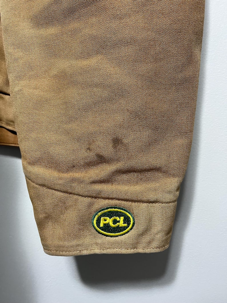 Puede incluir: Primer plano de una manga de chaqueta de trabajo color canela. La manga tiene una tela texturizada y un peque&ntilde;o parche bordado con las letras "PCL" en blanco, rodeado por un &oacute;valo verde con borde amarillo.