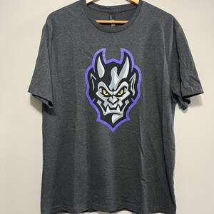 May include: A charcoal gray short-sleeve t-shirt featuring a graphic print of a stylized demon head with yellow eyes, gray skin, and sharp teeth, outlined in a vibrant purple border. The shirt is displayed on a wooden hanger with a small tag.