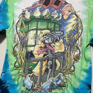 May include: Tie-dye t-shirt with a colorful graphic. The design features a skeleton in a yellow coat looking through a telescope, surrounded by birds and a lightning bolt. The shirt has a white base with blue and green tie-dye.