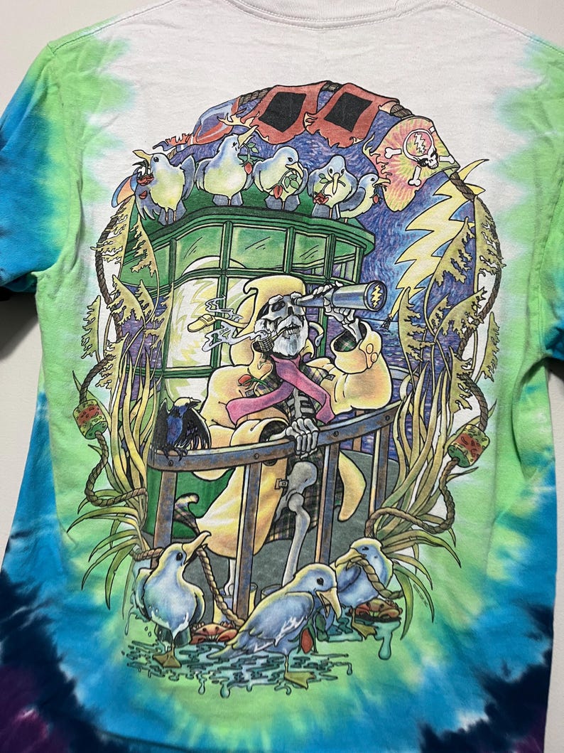 May include: Tie-dye t-shirt with a colorful graphic. The design features a skeleton in a yellow coat looking through a telescope, surrounded by birds and a lightning bolt. The shirt has a white base with blue and green tie-dye.