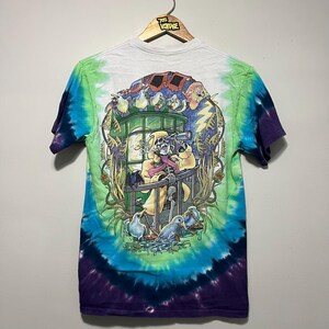 May include: A tie-dye t-shirt with a colorful graphic design featuring a skeleton, birds, and a train. The shirt has a white top, green and blue swirls, and a purple bottom. The shirt is hanging on a wooden hanger.