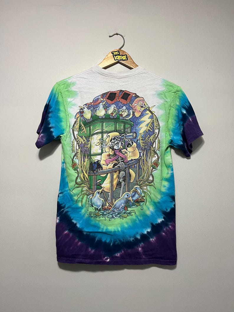 May include: A tie-dye t-shirt with a colorful graphic design featuring a skeleton, birds, and a train. The shirt has a white top, green and blue swirls, and a purple bottom. The shirt is hanging on a wooden hanger.