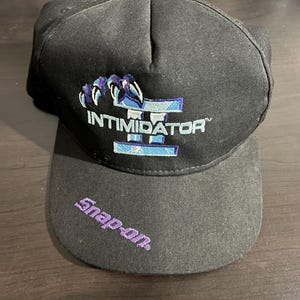 Peut inclure: Casquette de baseball noire avec le mot "INTIMIDATOR" en blanc et bleu, et un motif de griffes. Le bord est brodé du mot "Snap-on" en violet.