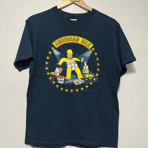 May include: Navy blue t-shirt featuring a cartoon image of Homer Simpson in a white diaper, surrounded by food and money, under the words "AMERICAN IDLE" in a yellow banner. The design is encircled by yellow stars.
