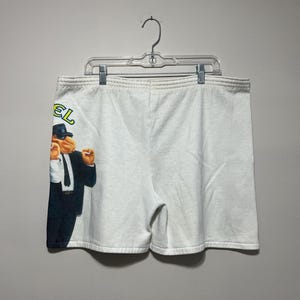 Pu&ograve; includere: Pantaloncini sportivi bianchi con una grafica di un cammello in giacca e cravatta e la scritta "Camel" in giallo e verde. I pantaloncini sono appesi a un'appendiabiti di plastica trasparente.