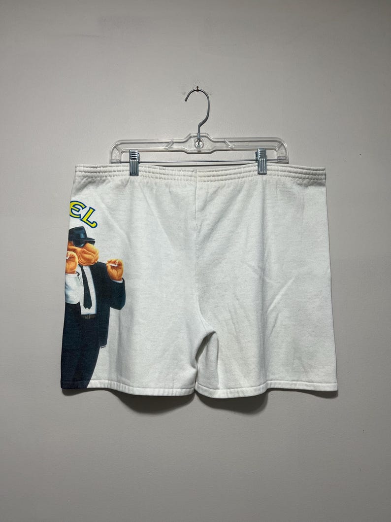 Pu&ograve; includere: Pantaloncini sportivi bianchi con una grafica di un cammello in giacca e cravatta e la scritta "Camel" in giallo e verde. I pantaloncini sono appesi a un'appendiabiti di plastica trasparente.
