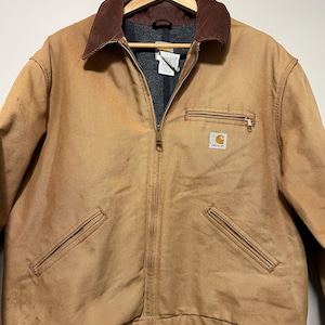 Puede incluir: Chaqueta de trabajo Carhartt marr&oacute;n con cuello de pana marr&oacute;n y forro interior gris. La chaqueta tiene cierre de cremallera, dos bolsillos delanteros y un bolsillo en el pecho con el logotipo de Carhartt. La chaqueta est&aacute; colgada en una percha de madera.