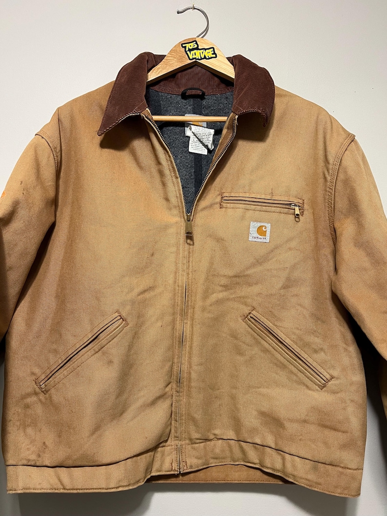 Puede incluir: Chaqueta de trabajo Carhartt marr&oacute;n con cuello de pana marr&oacute;n y forro interior gris. La chaqueta tiene cierre de cremallera, dos bolsillos delanteros y un bolsillo en el pecho con el logotipo de Carhartt. La chaqueta est&aacute; colgada en una percha de madera.