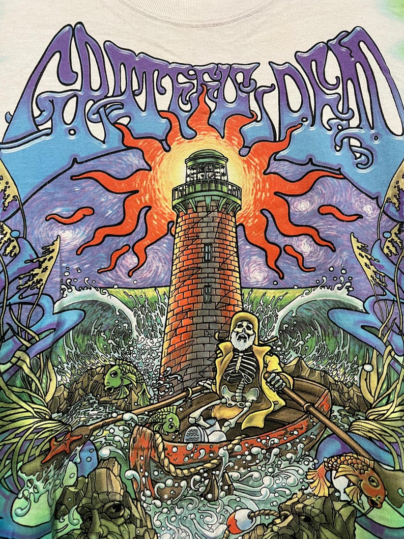 May include: White t-shirt featuring a colorful graphic design. The design includes a lighthouse, a skeleton in a boat, and the words "Grateful Dead" in a swirling font. The artwork is vibrant with blues, oranges, and greens.