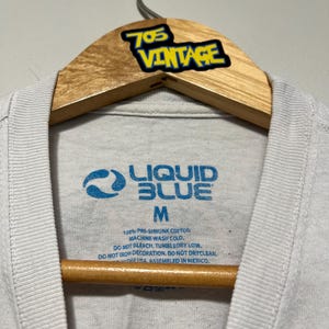 May include: Close-up of a white t-shirt with a Liquid Blue logo in blue. The shirt is size M and made of 100% pre-shrunk cotton. The tag also includes washing instructions. The shirt is hanging on a wooden hanger with a "705 Vintage" sticker.