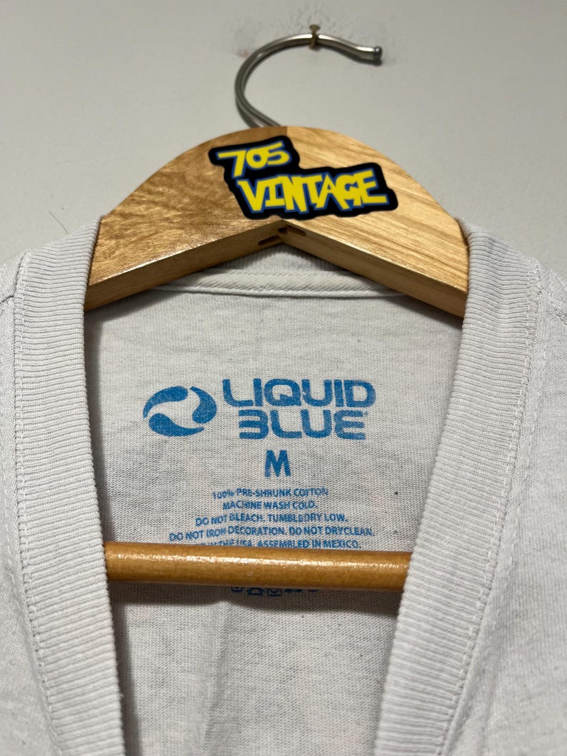 May include: Close-up of a white t-shirt with a Liquid Blue logo in blue. The shirt is size M and made of 100% pre-shrunk cotton. The tag also includes washing instructions. The shirt is hanging on a wooden hanger with a "705 Vintage" sticker.