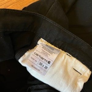 May include: A white tag with black text that reads "carhartt" and "RELAXED FIT" with the size "38 x 28" and the style number "102291 001". The tag is attached to a pair of black trousers.