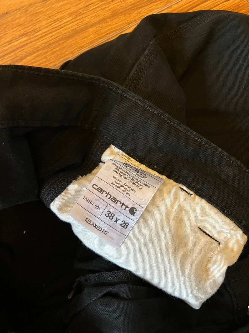 May include: A white tag with black text that reads "carhartt" and "RELAXED FIT" with the size "38 x 28" and the style number "102291 001". The tag is attached to a pair of black trousers.