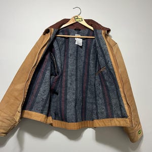 Puede incluir: Chaqueta de trabajo marr&oacute;n con cuello de pana marr&oacute;n, colgada de una percha de madera. El interior de la chaqueta presenta un forro a rayas grises y rojas. La chaqueta tiene cierre de cremallera y un peque&ntilde;o logotipo verde en la manga.