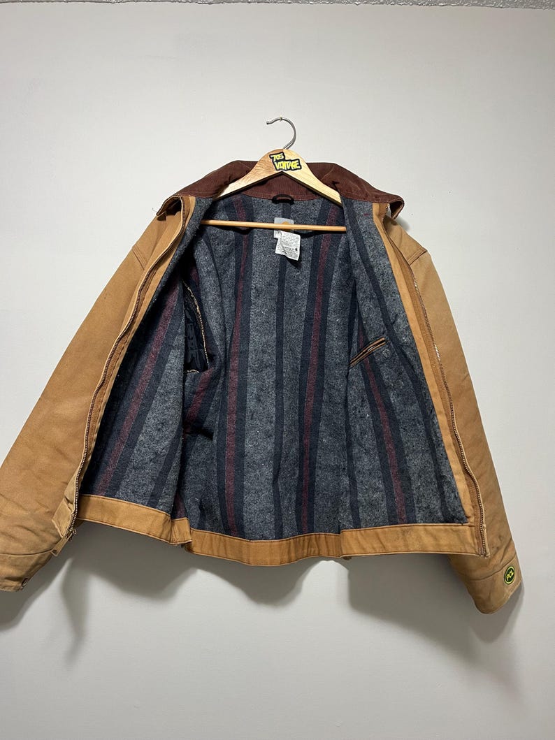 Puede incluir: Chaqueta de trabajo marr&oacute;n con cuello de pana marr&oacute;n, colgada de una percha de madera. El interior de la chaqueta presenta un forro a rayas grises y rojas. La chaqueta tiene cierre de cremallera y un peque&ntilde;o logotipo verde en la manga.