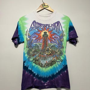 May include: Tie-dye t-shirt with a graphic featuring a lighthouse, sun, and text "Grateful Dead". The shirt has a white collar and sleeves, with a blue, green, and purple tie-dye pattern. The shirt is hanging on a wooden hanger.
