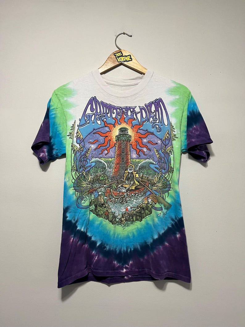 May include: Tie-dye t-shirt with a graphic featuring a lighthouse, sun, and text "Grateful Dead". The shirt has a white collar and sleeves, with a blue, green, and purple tie-dye pattern. The shirt is hanging on a wooden hanger.